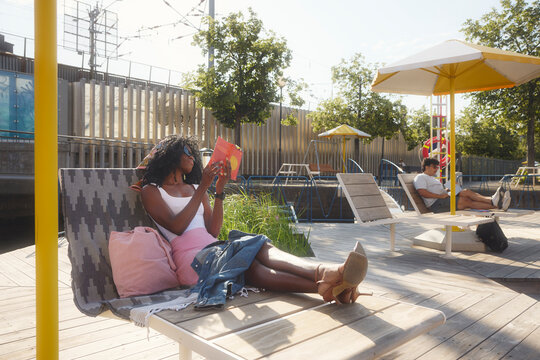 Carefree woman reading book while sitting on deck chair at sunny day