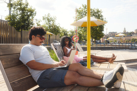 Man using laptop while sitting with female friend on deck chair at sunny day