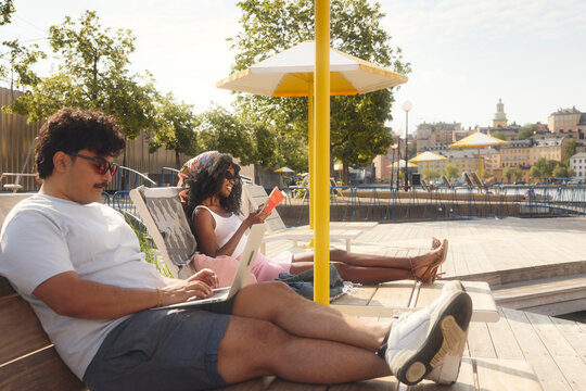 Man using laptop sitting on deck chair beside female friend reading book at sunny day