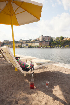 Juice bottle kept on sand near deck chair with book and bag under beach umbrella