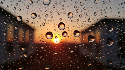 Mesmerizing close-up of raindrops clinging to a window pane, perfectly framing a blurred, warm sunset between residential buildings, creating a serene and reflective atmosphere