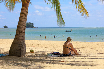 Blonde woman in bikini sitting on a sand under the palm tree, vacation on tropical sea beach