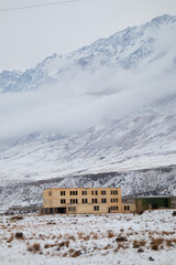 Fototapeta premium A derelict multi-story building standing in a snowy field with low clouds covering the massive mountain range behind it