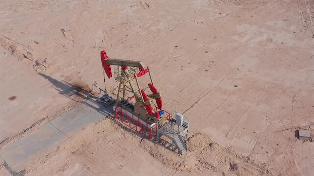 Brightly painted oil pump jack with red counterweights and beige structure operating in desert oilfield. Drone view, industrial equipment contrasting with dry barren soil and clear horizon.
