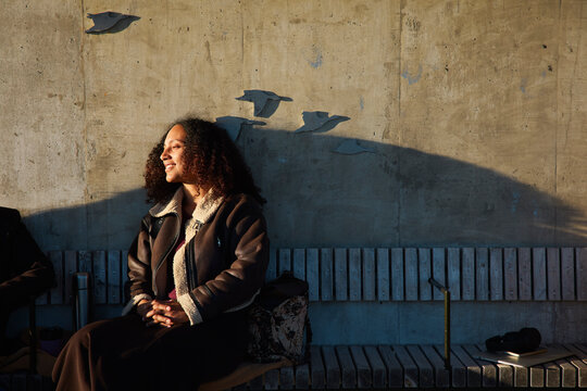Smiling woman in leather jacket sitting on bench against wall at par