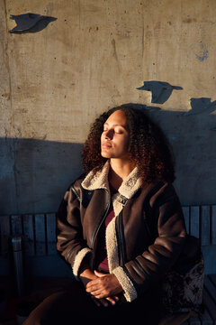 Woman in leather jacket sitting with eyes closed on bench at park