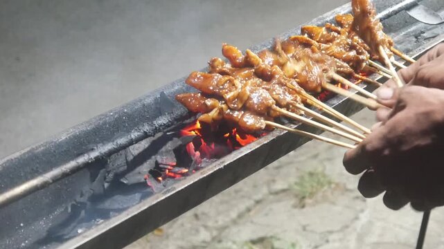 The process of grilling chicken satay over hot coals on a long grill. Rows of skewers are golden brown with a glistening of spices, while flames and smoke rise from the grill.