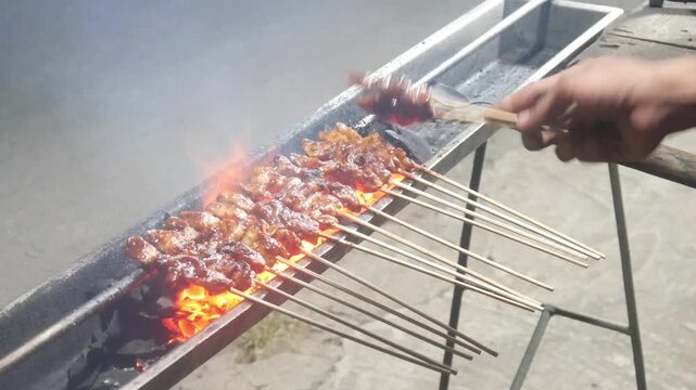 The process of grilling chicken satay over hot coals on a long grill. Rows of skewers are golden brown with a glistening of spices, while flames and smoke rise from the grill.