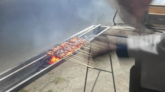 The process of grilling chicken satay over hot coals on a long grill. Rows of skewers are golden brown with a glistening of spices, while flames and smoke rise from the grill.