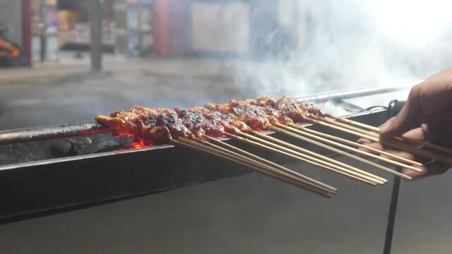 The process of grilling chicken satay over hot coals on a long grill. Rows of skewers are golden brown with a glistening of spices, while flames and smoke rise from the grill.