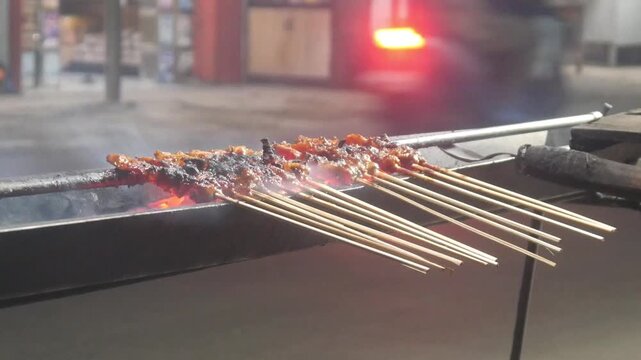 The process of grilling chicken satay over hot coals on a long grill. Rows of skewers are golden brown with a glistening of spices, while flames and smoke rise from the grill.