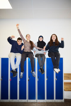 Full length of cheerful friends enjoying while sitting on locker in school