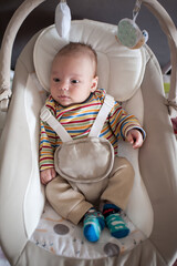 A cute baby with a striped shirt sits secured in a baby swing, looking off to the side. Toys hang above the swing, providing visual stimulation for the infant