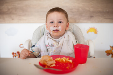 A young boy sits in a high chair with food on his face and bib. He is eating lunch at home. A red plate with food and a red cup are on the table in front of him