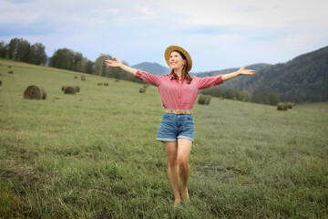 A woman wearing a straw hat and a red plaid shirt stands in a green field with rolls of hay, her arms spread out and her face lit up with a smile, surrounded by hills and a forest. © ELENA