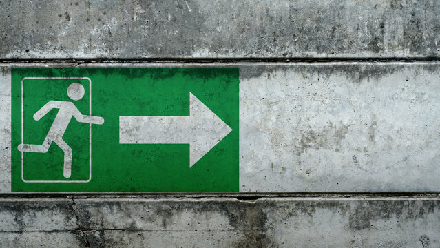 Green emergency exit sign with a running man and arrow, painted on a textured concrete wall. The sign indicates the direction of the nearest emergency exit route. Concept of Evacuation