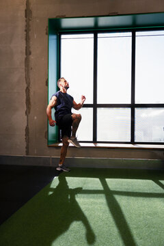 Full length of active man doing high knee exercise near window in gym