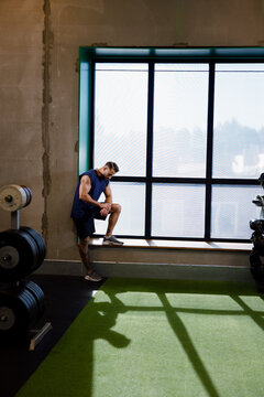 Full length of exhausted man standing near window while taking break after warm up exercise in gym