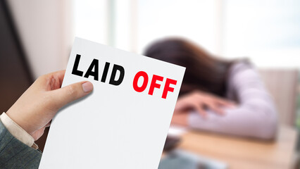 Hand holding a layoff notice with the words LAID OFF printed on it. In the background, a despondent employee rests their head on a desk, highlighting the emotional impact of job loss. Concept of Laid 