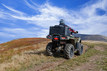 A person riding a ATV with a black cargo box drives up a dry, grassy dirt path toward rolling hills under a dramatic blue sky with wispy white clouds.