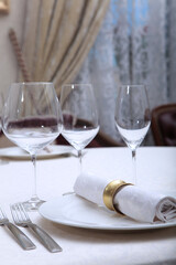 A white napkin with a ring lies on a plate. Festive table setting. A napkin ring. Wine glasses on the table. White plates. VIP lounge. White tablecloth with textured pattern. Part of the interior.