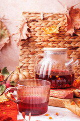 Glass cup with red herbal tea and teapot in background on rustic table with sea buckthorn, apple and dry leaves
