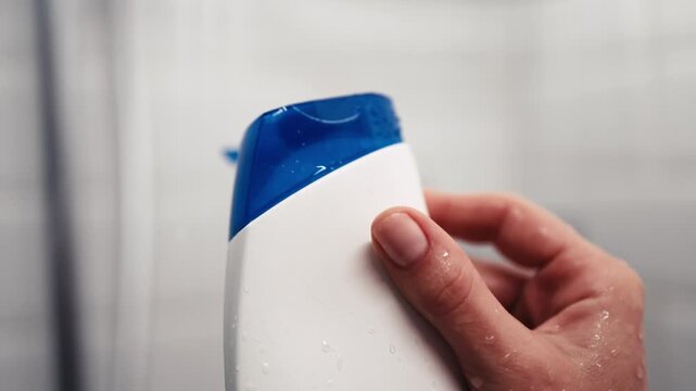 Hand holds a shampoo bottle in the shower, capturing the moment of opening with water droplets enhancing the scene. Self-care concept and soothing bathroom atmosphere.