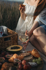 Woman’s Hand Holding a Glass of White Wine at a Picnic with Exotic Fruits, Croissants, and Grapes in the Grass