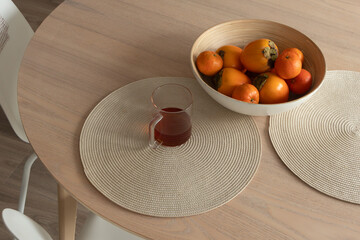 Glass cup of hot tea and bowl of fresh persimmons and mandarins on wooden dining table. Cozy home atmosphere, modern minimalist kitchen interior