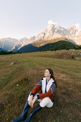 Naklejka premium woman sitting on meadow in mountain landscape, nature scene with relaxed pose and closed eyes, outdoors lifestyle with warm jacket and boots enjoying fresh air and peaceful view