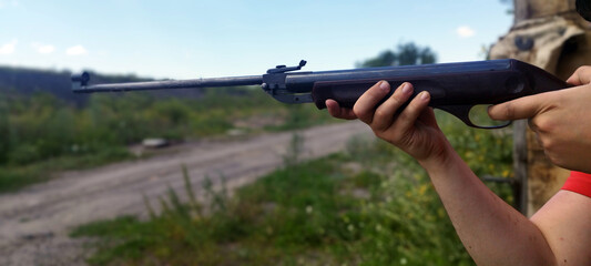 A person aims an air rifle outdoors, holding it steady with both hands. The background shows a dirt path and lush green field under a bright blue sky