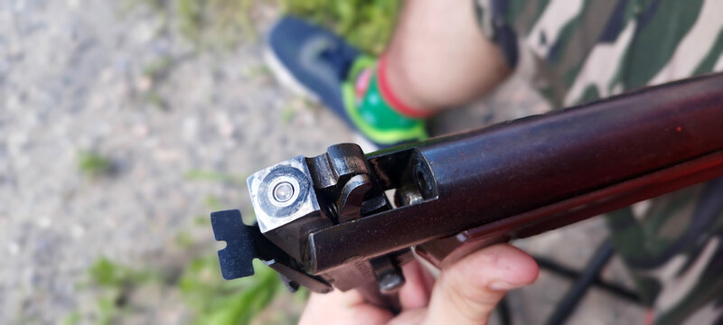A close-up of an air rifle's bolt, showing its mechanics. The man holding the rifle is wearing camouflage, and the outdoor setting is brightly lit.