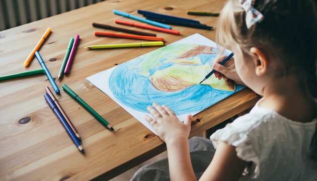 Over-the-shoulder view of a child drawing a colorful Earth illustration on a large sheet of paper, surrounded by vibrant colored pencils on a warm wooden desk with soft natural lighting, pastel and br