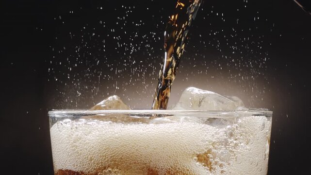 Close-up shot of glass top and cola drink being poured into a glass. Visible fizzy splash and rising carbonate bubble close up on dark background.