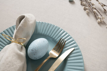 Elegant Easter table setting with blue speckled egg, gold cutlery and linen napkin on pastel plate and linen tablecloth. Soft spring decor and holiday atmosphere.