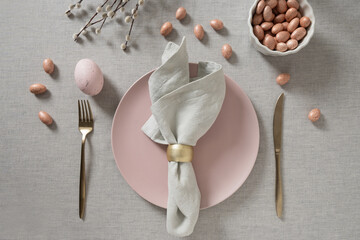 Stylish Easter table setting with pastel pink egg, gold cutlery and willow branches on soft linen background. Spring holiday decor.