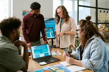 Diverse group of young adult men and women collaborating around table, discussing digital project with laptops and color samples, Caucasian woman presenting smartphone screen to team