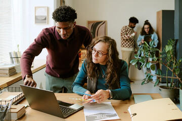 Young adult Black man standing beside young adult Caucasian woman working together at desk with...
