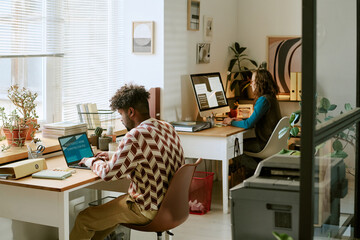 Young adult Black man working on laptop at desk in modern office while young Caucasian woman using desktop computer at workstation, both focused on tasks, surrounded by plants and office equipment