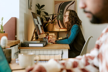 Caucasian young adult woman working at computer desk in modern office, focusing on screen while typing, with blurred Caucasian man in foreground using laptop, creative workspace setting
