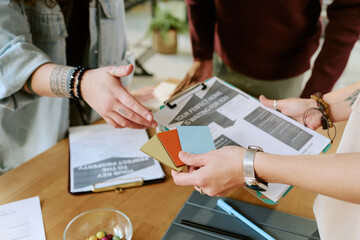 Caucasian woman and Black man collaborating at desk, reviewing color swatches and documents, hands visible exchanging samples, young adults discussing project ideas together