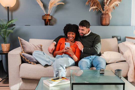 Happy couple sharing a moment while using a smartphone to explore an app on their cozy couch together, enjoying their time in a warm and inviting home atmosphere filled with love and connection. - Powered by Adobe