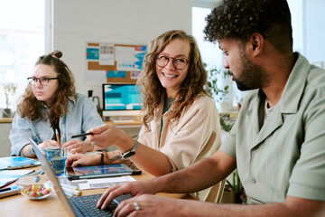 Caucasian young adult woman smiling and interacting with Black young adult man while working at desk with laptop, another Caucasian young adult woman focusing on computer in background