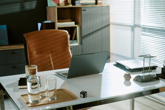 Modern office workspace featuring closed laptop, glasses of water, coffee cup, documents, and office supplies on desk with sunlight streaming through window blinds in background