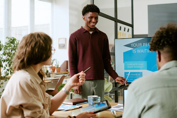 Young Black man standing and presenting information on digital screen to diverse group of young adults sitting at table, engaging in business meeting or collaborative discussion in office
