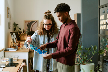 Caucasian young adult woman and Black young adult man standing together reviewing printed document in modern office setting, both focused on analyzing information and collaborating