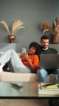 A joyful couple engages with their smartphone and laptop, sharing moments and experiences in a comfortable living space while relaxing together on a couch, embodying connection and togetherness.