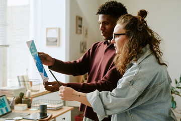 Young adult Black man and young adult Caucasian woman collaborating while holding and discussing printed charts in modern office setting, both focused on analyzing information together