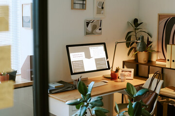 Modern home office workspace featuring desktop computer displaying real estate presentation, desk lamp, potted plants, organized shelves, framed abstract wall art, natural daylight streaming