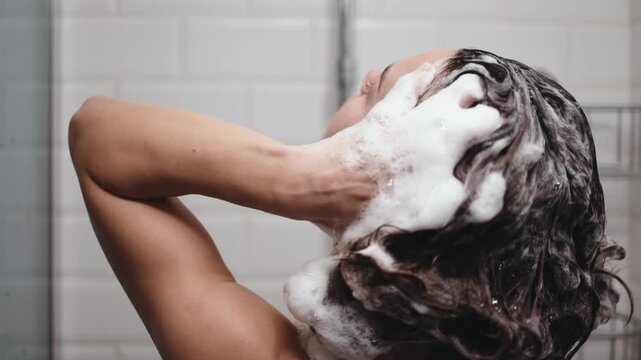 Woman with dark hair applies shampoo in the shower, highlighting the lathering process and hair texture. Hair care concept and self-care moment.